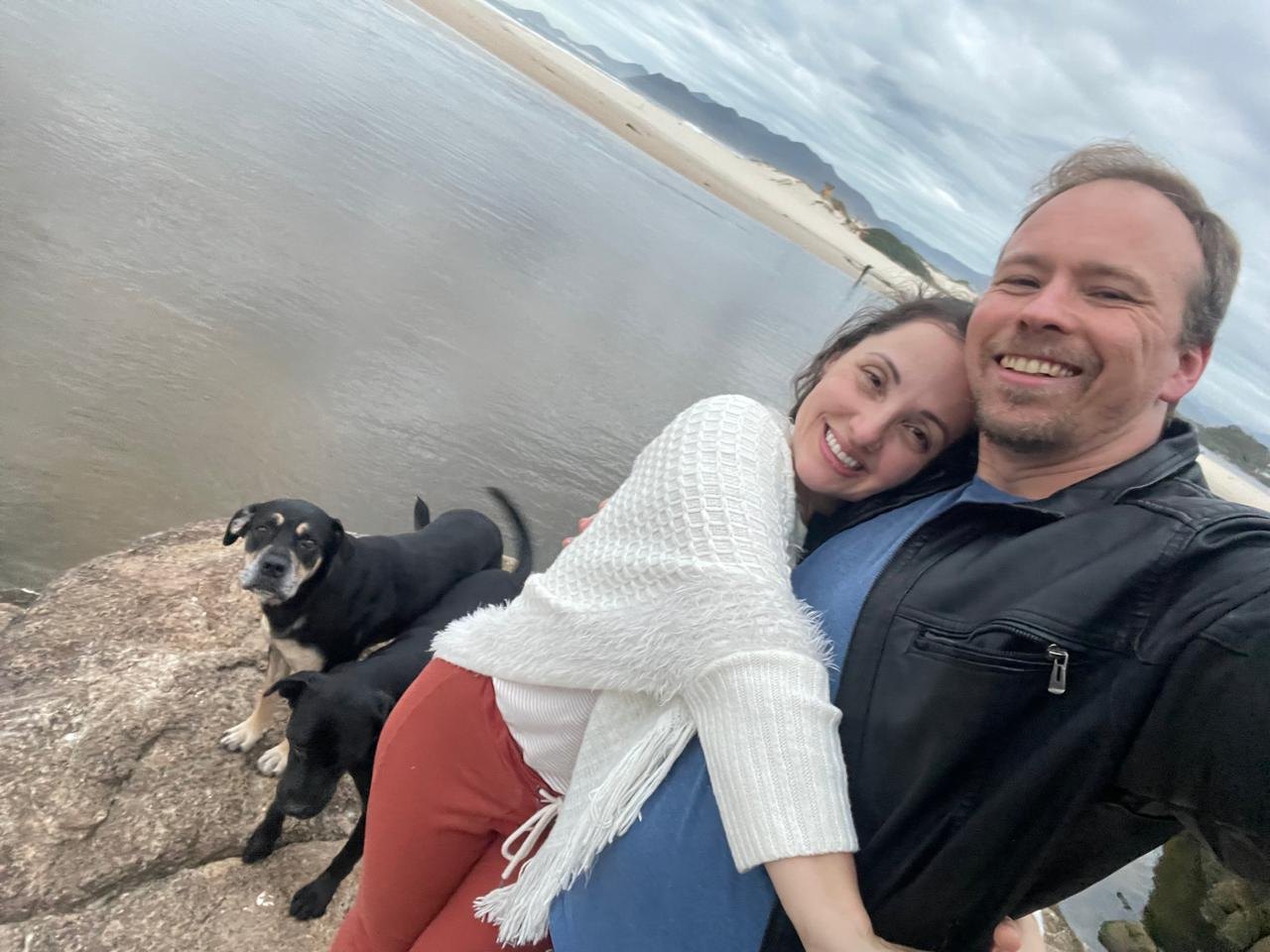Marcelo e Pâmela abraçados na praia, com os dois cachorros ao lado